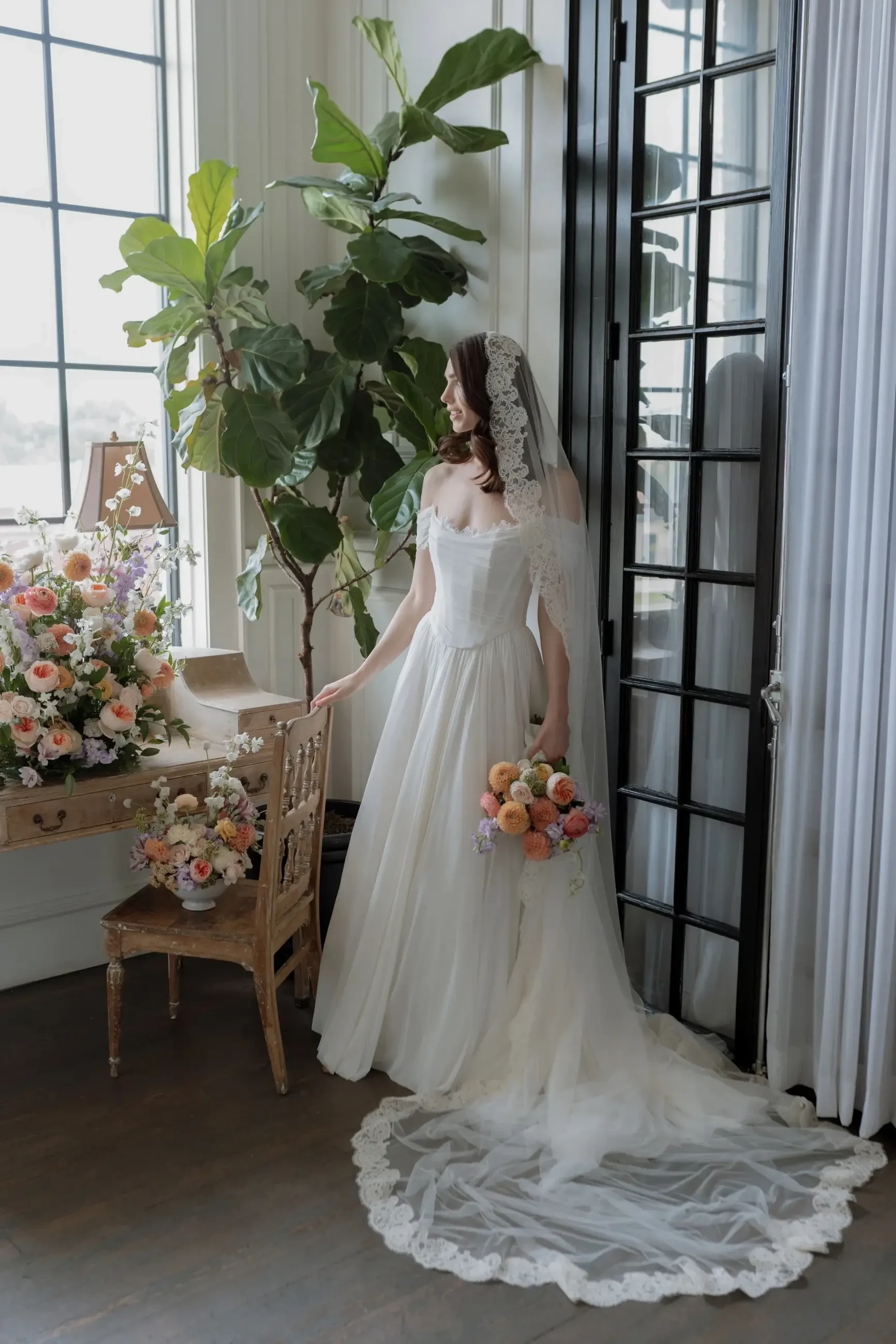A bride in a white wedding dress stands near a table adorned with flowers, holding a colorful bouquet. She has long hair and is wearing a veil, with large windows and greenery in the background.