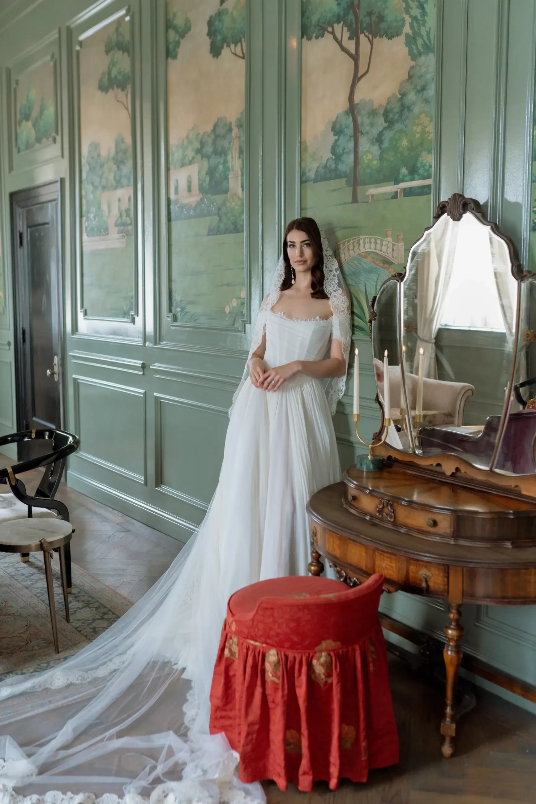 A woman in a white bridal gown stands beside a vintage vanity in an elegantly styled room with green walls and scenic wall art.