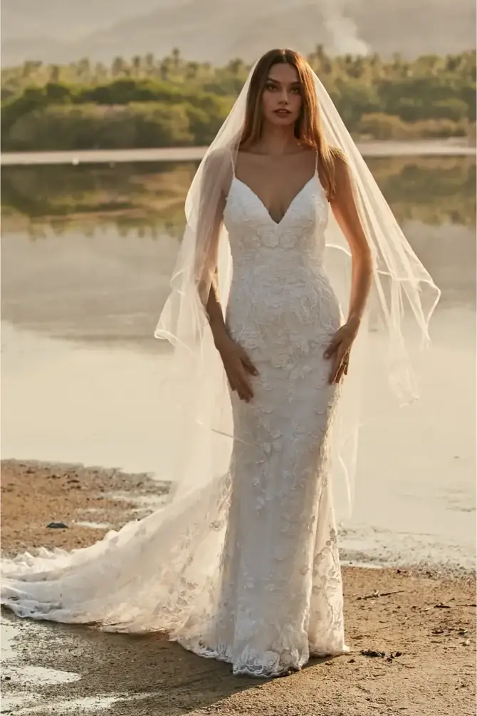 A woman in a white wedding dress stands near a body of water, wearing a long veil.
