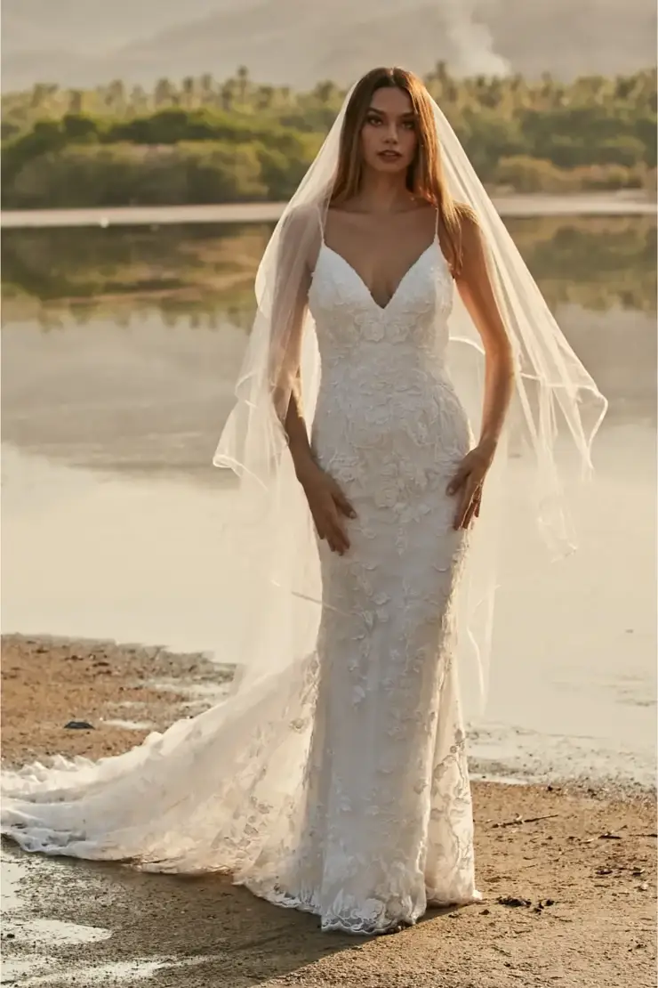 A bride wearing a beautiful white wedding dress with a deep V neckline and intricate floral details, standing by a serene body of water, with a soft veil flowing over her shoulders.