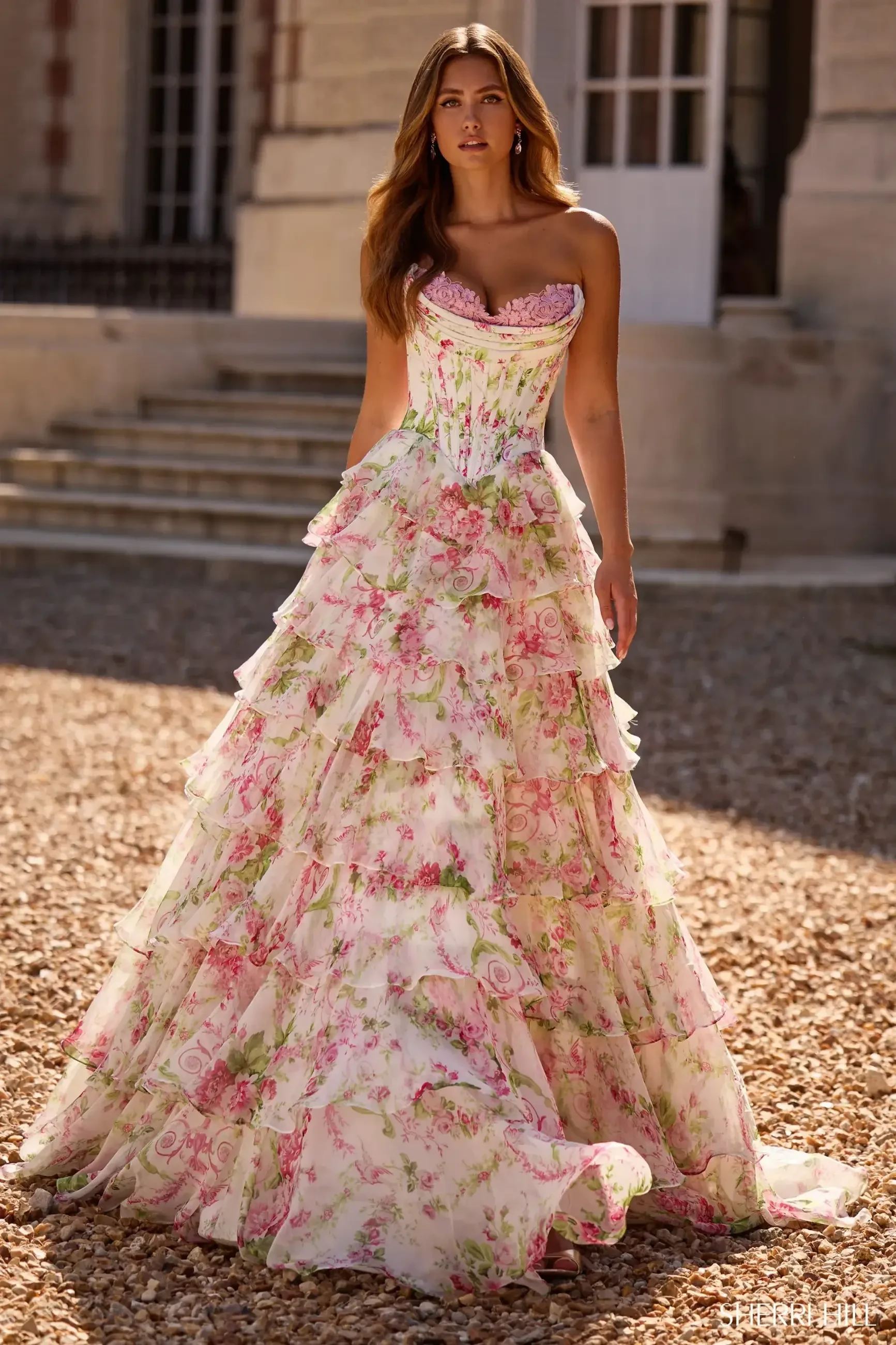 A woman wearing a floral strapless gown with layered ruffles, standing outdoors in front of a building.