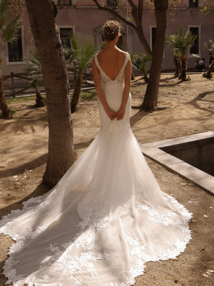 Bride in an elegant lace wedding dress stands outdoors, sunlight highlighting the train. Surrounded by trees in a serene, sandy courtyard.