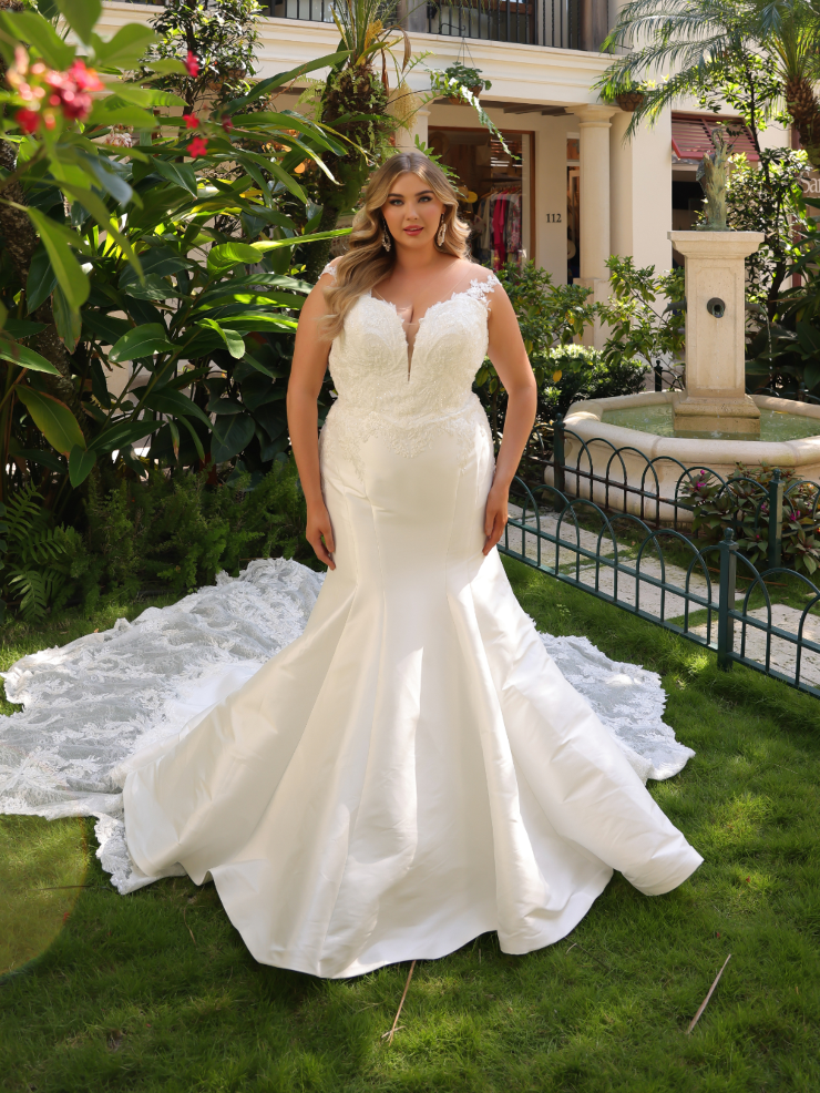 A woman in a formal, white wedding dress stands in a garden with lush greenery and a fountain behind her. The dress features intricate lace detail and a mermaid silhouette.