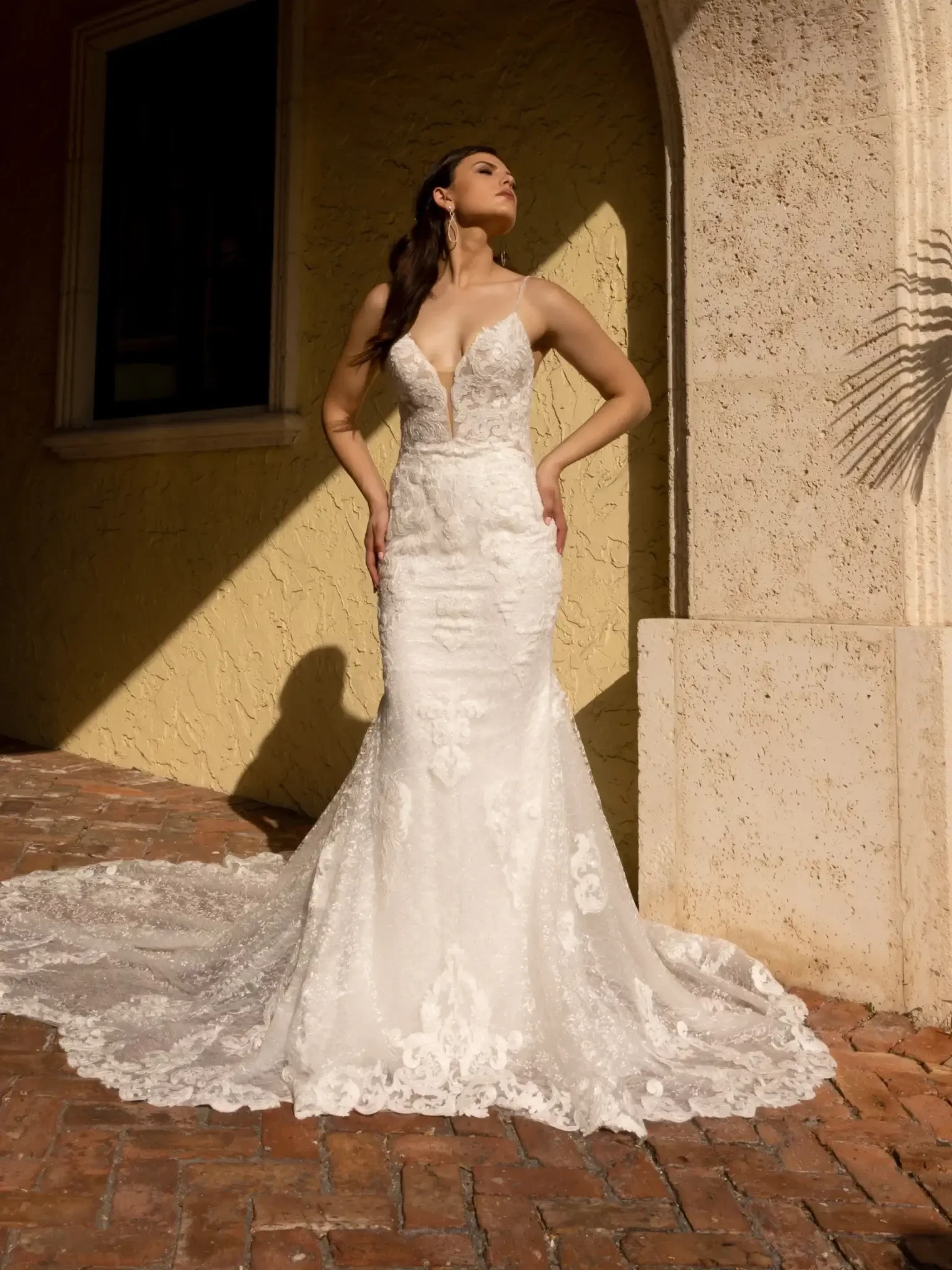 A woman in a lace mermaid wedding dress stands confidently in sunlight against a textured yellow wall. Her expression is serene and poised.