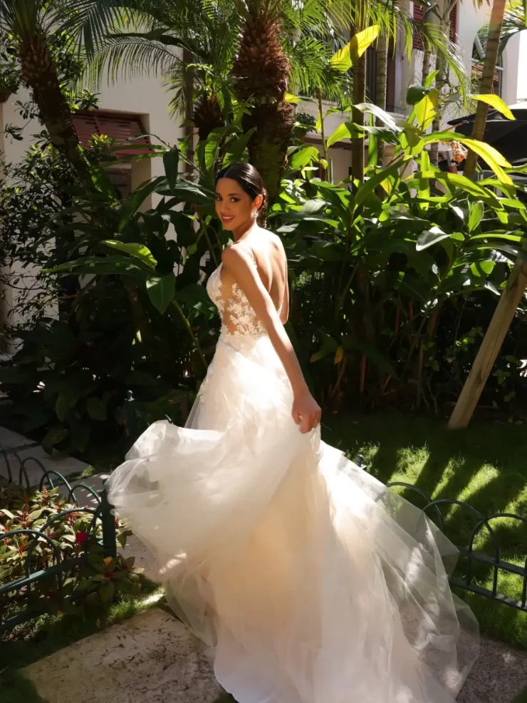 A woman in a flowing white wedding dress, featuring a lace back, is smiling and turning in a lush garden. The setting is vibrant and sunny.