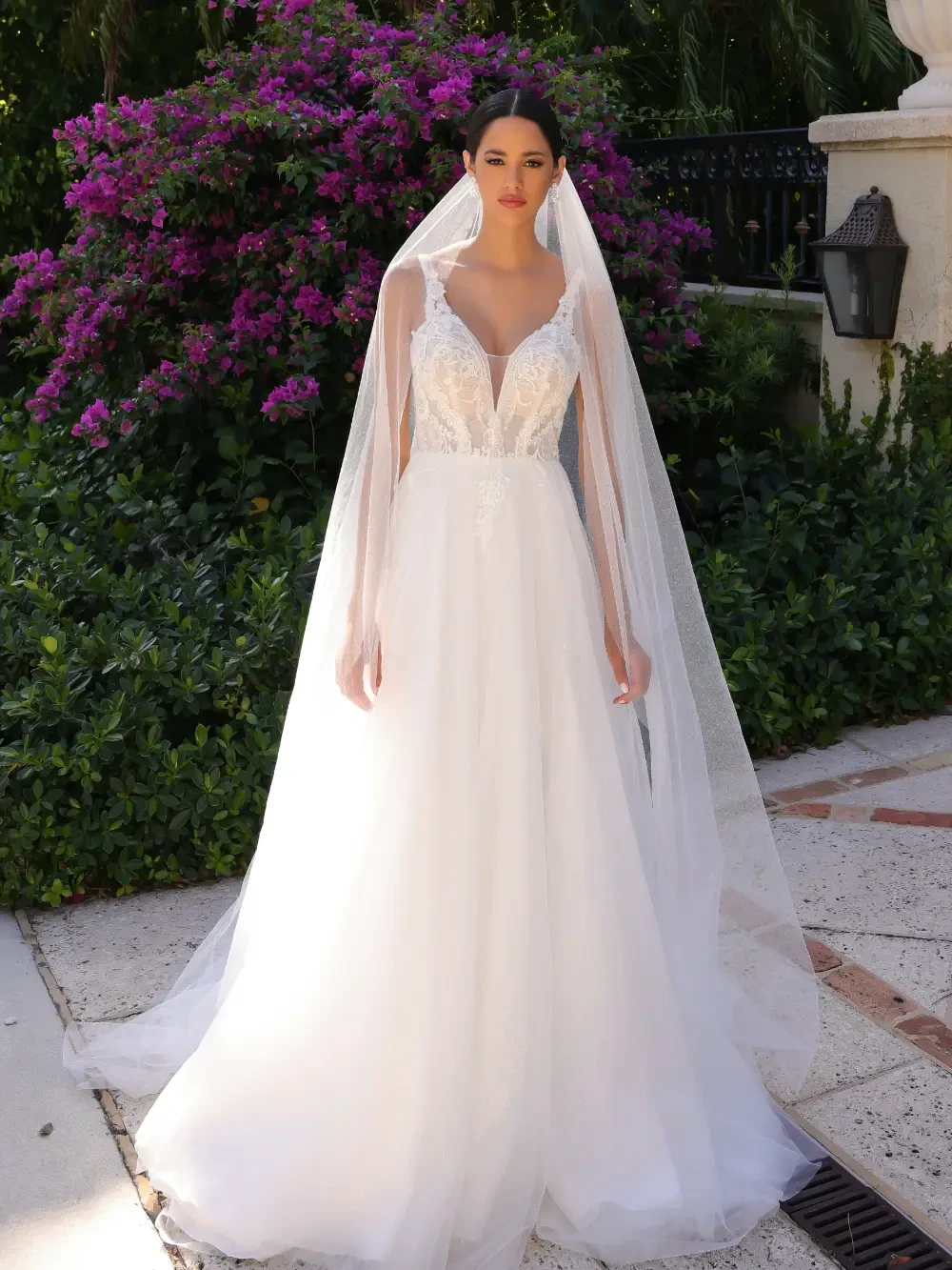 A bride wearing a beautiful white wedding dress with a lace bodice and a long veil, standing outdoors near purple flowers and greenery.
