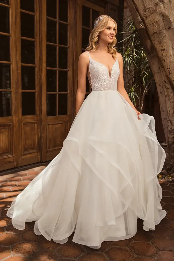 A woman in a flowing white wedding dress with a v-neckline and lace detailing, standing outdoors near wooden doors surrounded by greenery.