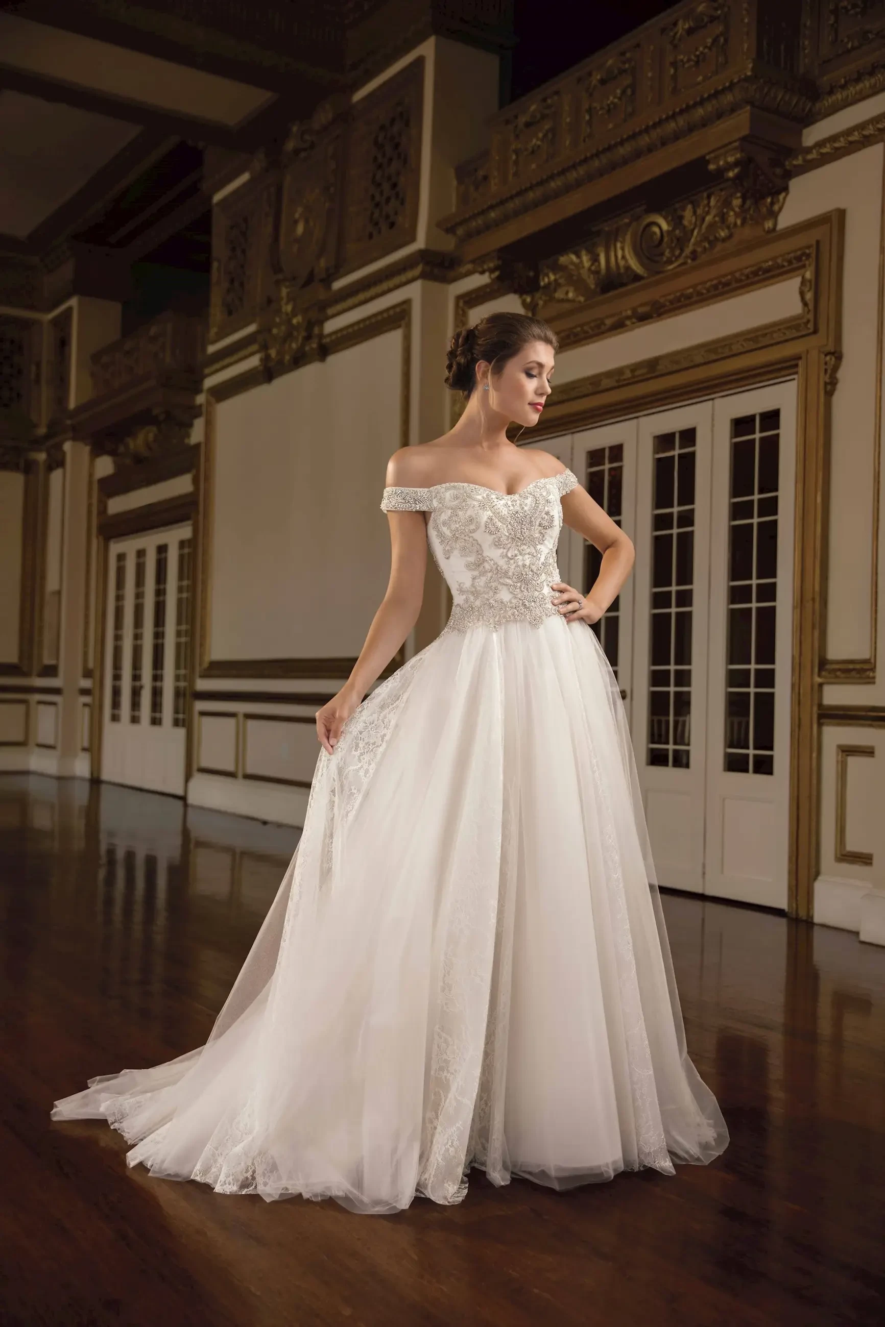 A model wearing an elegant off-the-shoulder wedding gown with intricate detailing, standing in a grand, well-lit interior space with wooden floors and decorative walls.