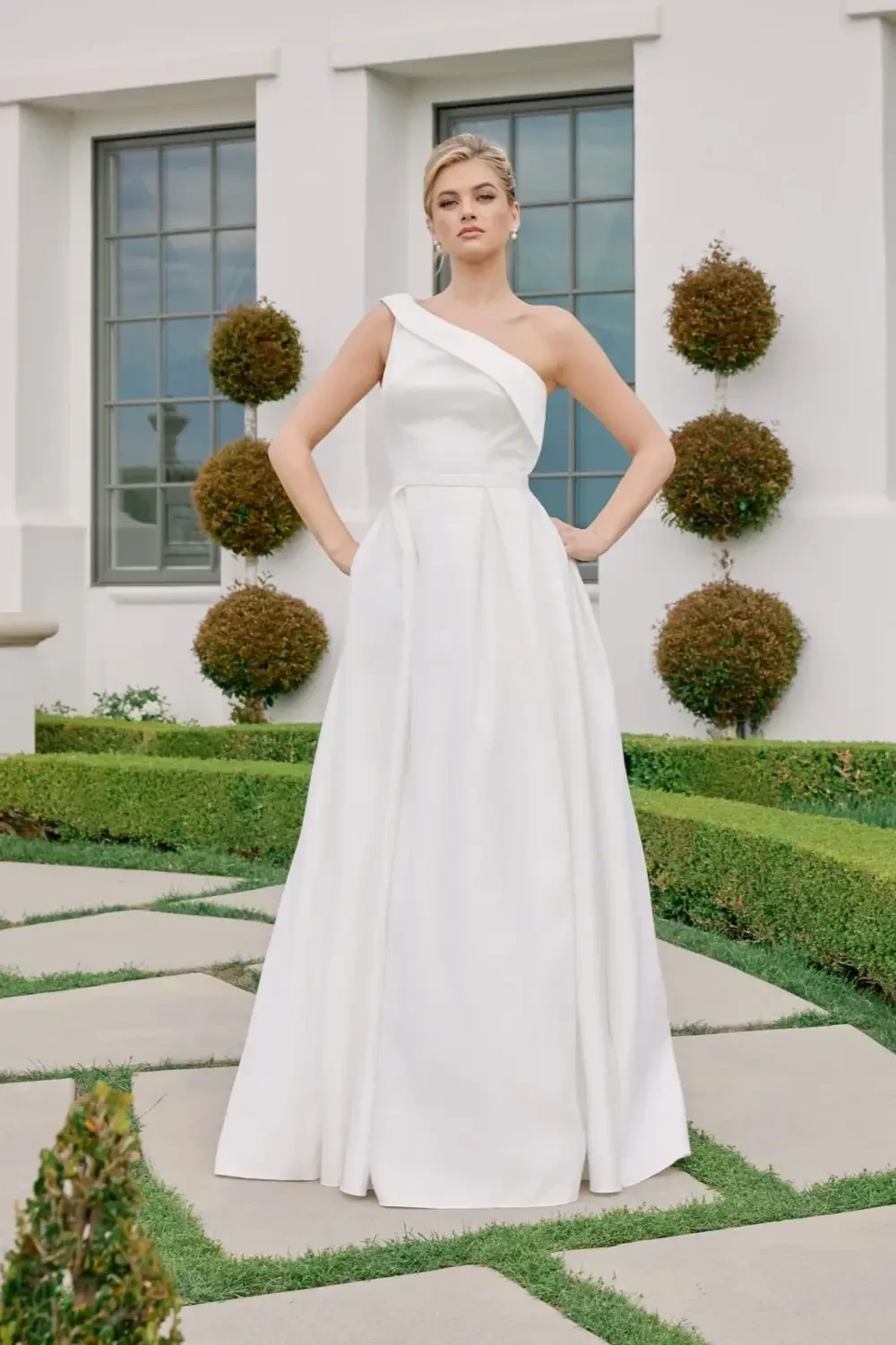 A woman in a stylish white gown with one shoulder exposed, standing confidently in a well-manicured garden.