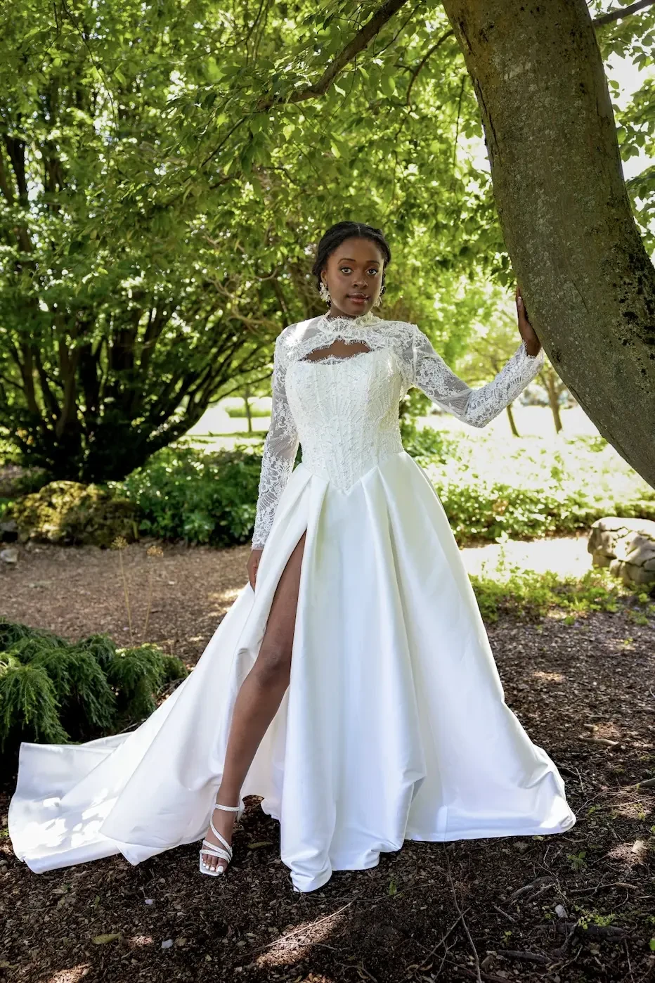 A person in a white wedding dress with a high slit and long lace sleeves poses outdoors beside a tree.