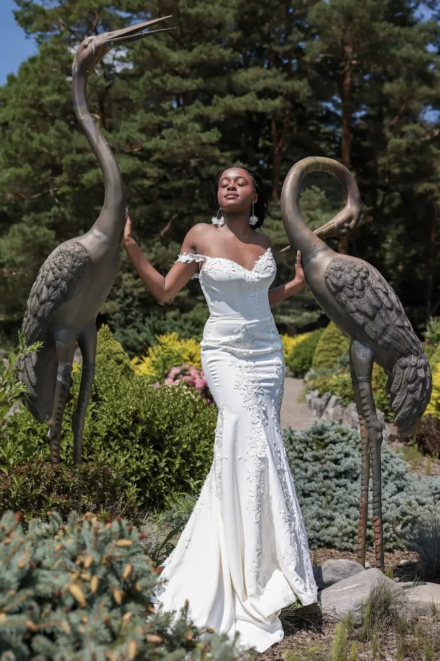 A woman in an elegant white gown poses between two statues of cranes in a lush garden.