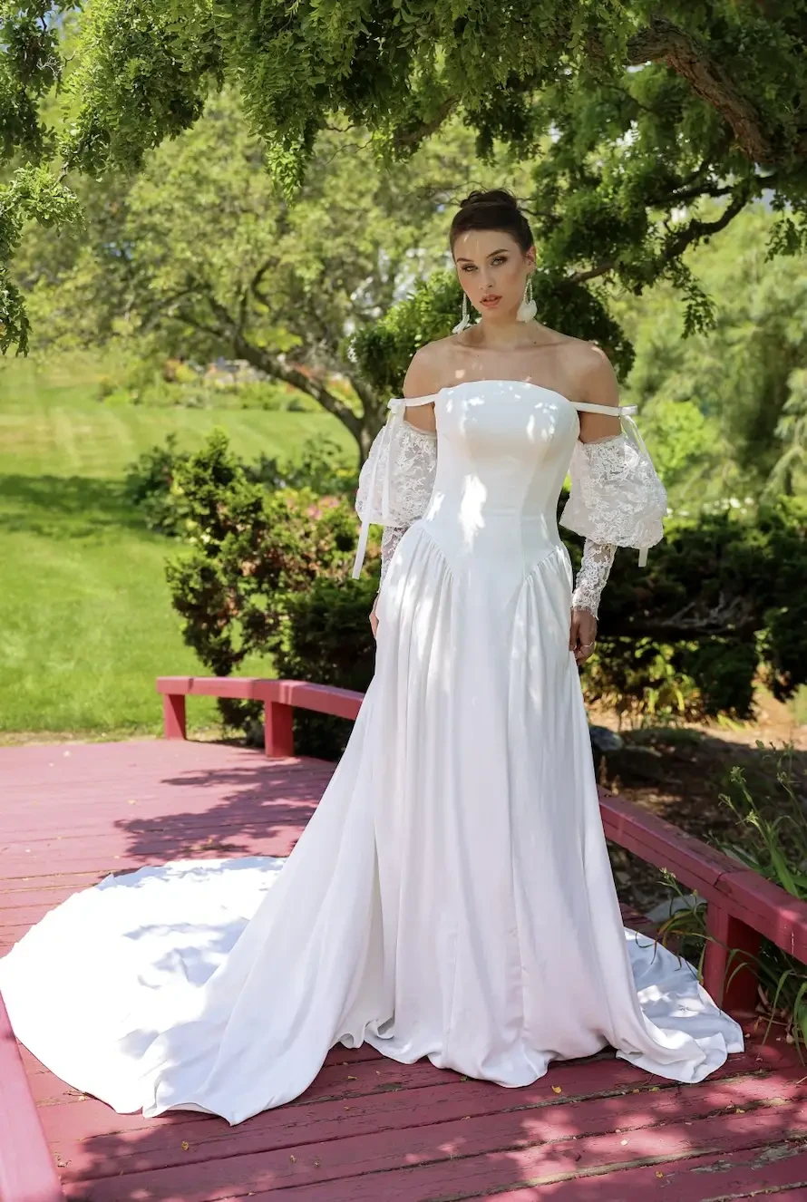 A woman in a white off-the-shoulder wedding dress stands on a pink wooden deck surrounded by greenery.