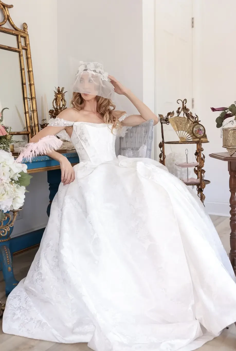 A bride wearing an elegant white wedding gown with lace details and a veil, sitting gracefully in a well-decorated room.