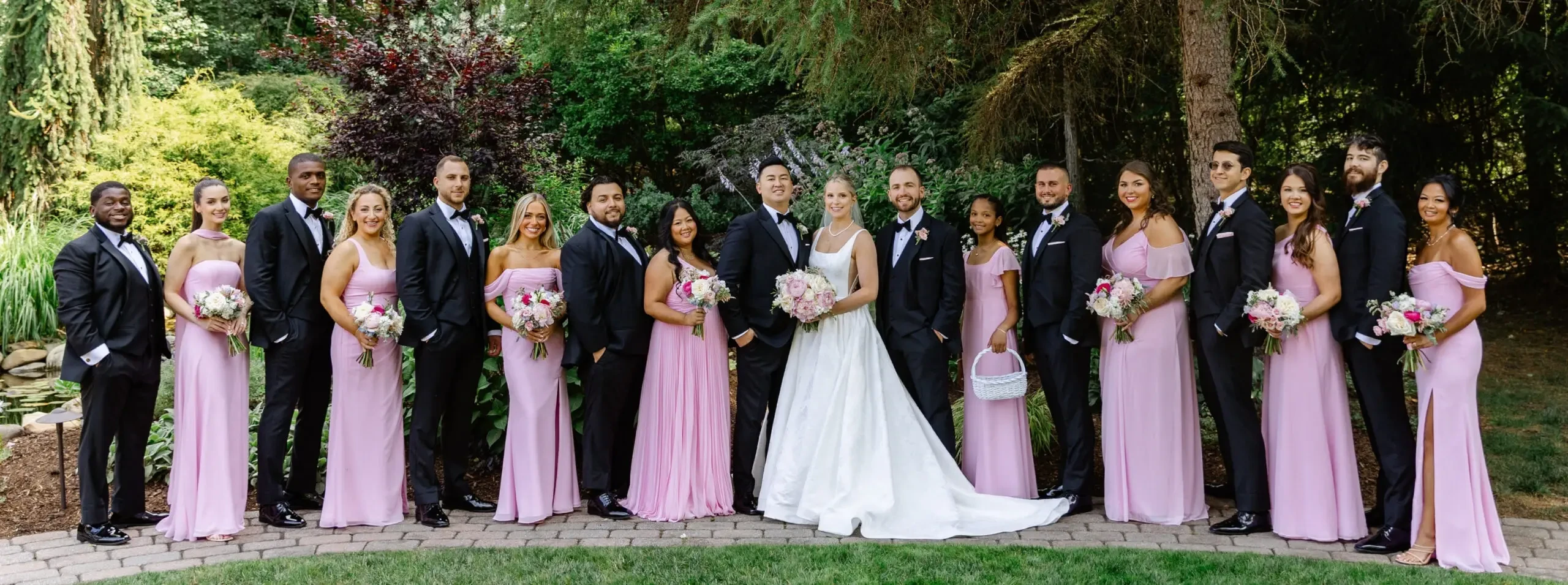 Bride and groom with their wedding party outdoors, featuring pink bridesmaid dresses and black tuxedos.